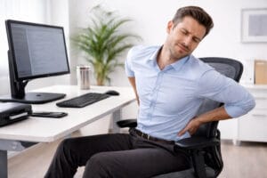 Hombre sentado en la oficina con dolor lumbar por mala ergonomía laboral y postura incorrecta