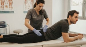 Fisioterapeuta de TMKine realizando una valoración del dolor en el glúteo al estar sentado a un paciente con ropa deportiva.
